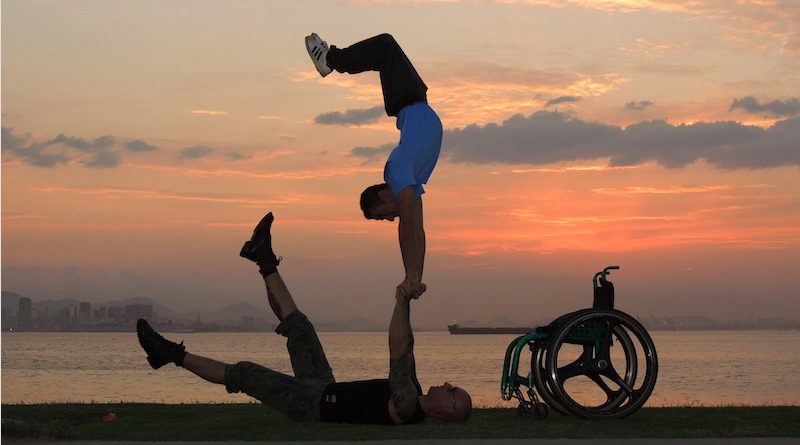 duo Dupla Mao Na Roda performing hand balancing by a lake at sunset. Photo description: There are bluish grey clouds and the sky is orange.Two handbalancers are silhoutted in the forefront. One hand balancer lays on the ground holding hands with the second hand balancer who is doing a handstand on the hands of the first. Also in the silhouette is the wheelchair of one of the handbalancers.