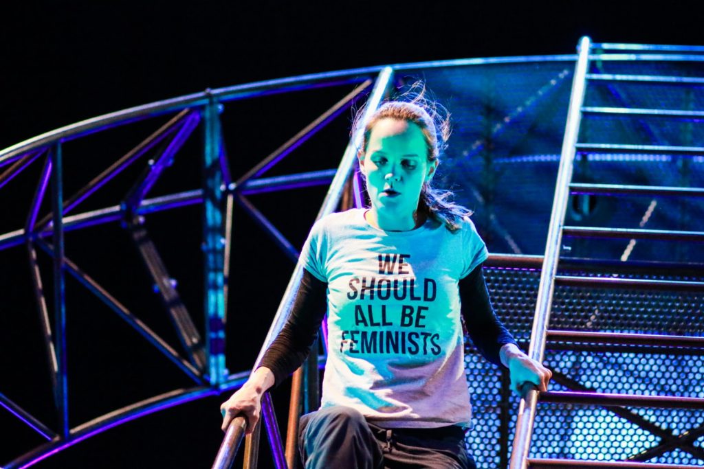 A white woman in her early thirties is climbing down the same steel structure. She is an aerialist and acrobat wearing a T shirt that says ‘ We should all be feminists’