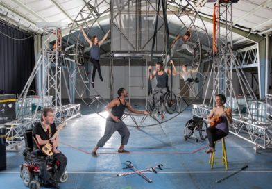 Seven performers disabled and non disabled surround a steel circular structure that they hang from . Two artists seated in front  play stringed unique instruments. Crutches lay on the floor . An artist who is a wheelchair user is suspended in his wheelchair from the structure.