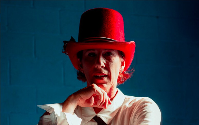 Aloysia Gavre staring into the camera thoughtfully, resting a hand on her chin, wearing a white button down and a large red hat