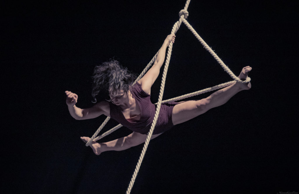 An aerial rope performer hangs from a suspended middle split