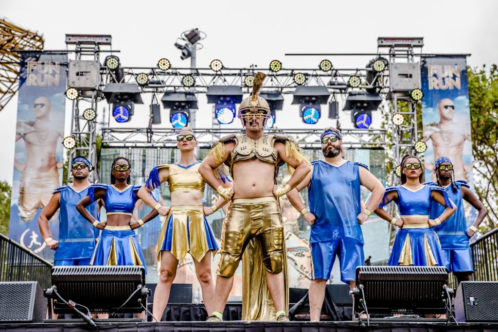 A group of performers stand on an outdoor stage, glad in blue and gold costumes