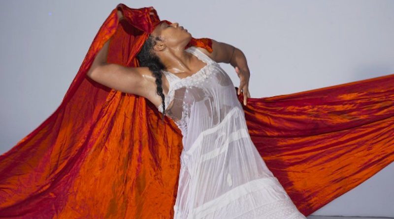 A Black woman wears a wet white gown, leaning back into a long crimson sash