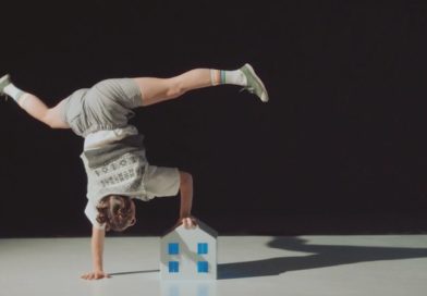 A handbalancer wearing a school uniform holds a one armed handstand, holding a small toy house and waving their legs