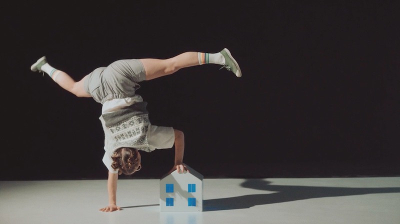 A handbalancer wearing a school uniform holds a one armed handstand, holding a small toy house and waving their legs