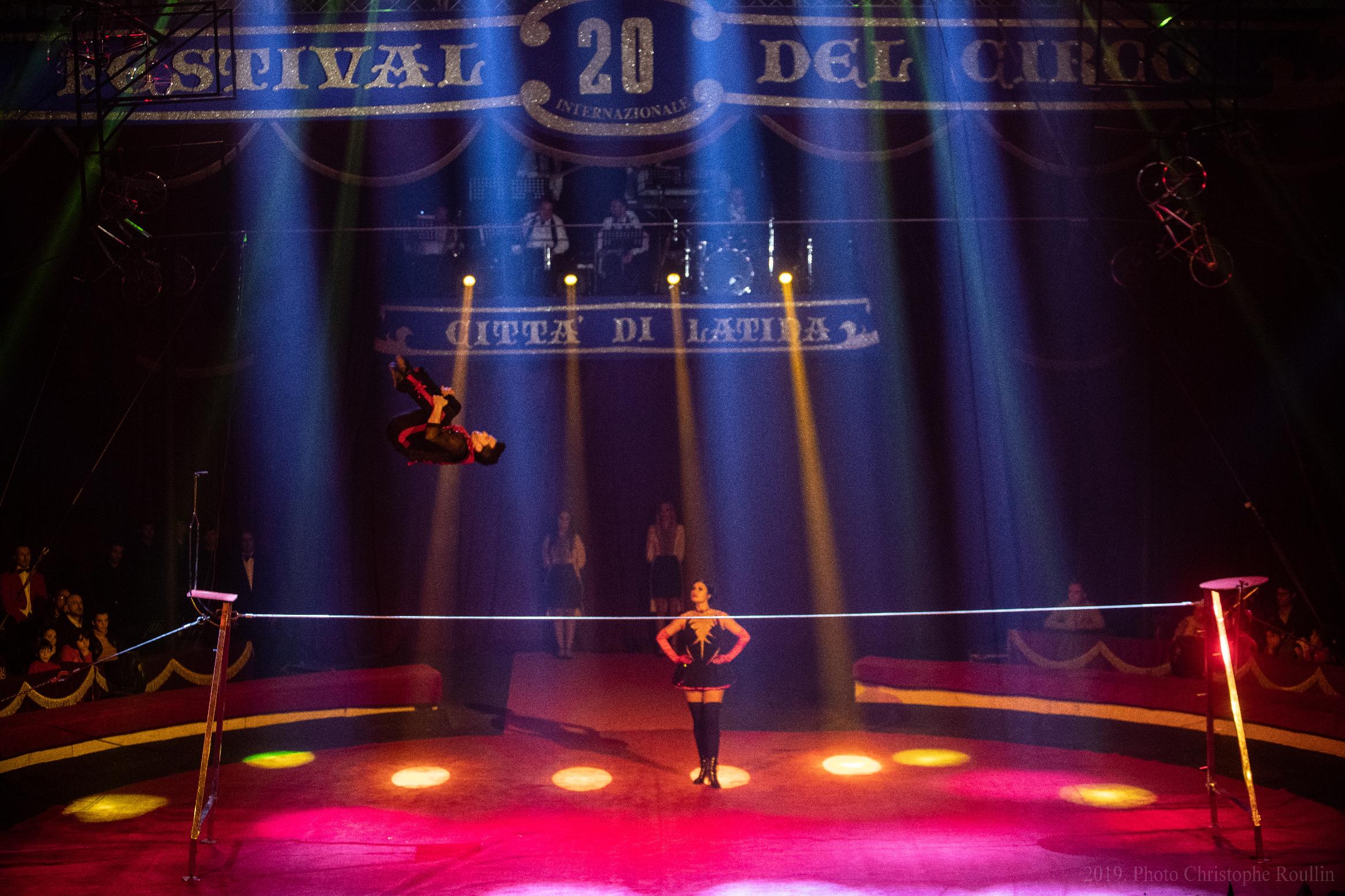 A performer on a stage lit in tones of red and purple does a back flip on the tight wire