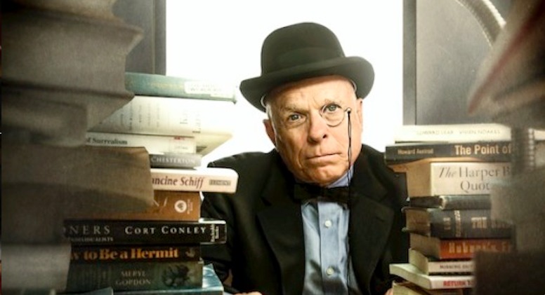 Jim Moore stares into the camera wearing spectacles and a black hat, surrounded by books