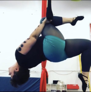 an aerialist hangs from one knee on a trapeze