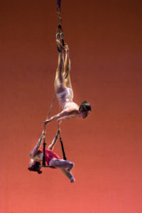 A mid-performance photo on stage of Vanessa Furlong and Erin Ball, both white. Two ropes with a bar between them at the bottom, a trapeze, hangs from the ceiling. Vanessa is upside down with her legs wrapped in the ropes, she arches, pushing her hands into the ropes. Below her, Erin, an amputee whose lower legs end just below the knees, sits on the bar and arches in the opposite direction of Vanessa. The lighting is orange. Vanessa’s outfit matches her skin and Erin wears a red bodysuit. 