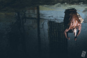 The image is upside down. Marco Motta faces away from the camera, seated on a blue-grey warehouse floor. The back wall is textured and darker. His wrists are looped into aerial straps. His back musculature is pronounced. The image has a watery quality.