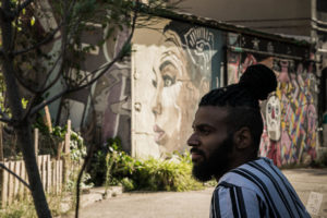 In the background a mural of a face is surrounded by greenery. In the foreground, Marco Motta is profiled looking to the left wearing a striped shirt.