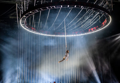 an aerialist flies beneath circular scaffolding with a back drop of ropes