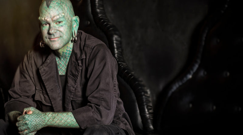 a male circus sideshow performer with lizard body modifications is seated in a dark robe against a dark background