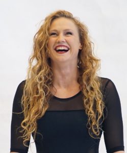Rowan Hayden-White stands smiling. Her curly blonde hair falls around her shoulders. She is wearing a long sleeved black top