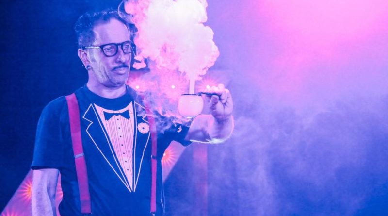 a male sideshow circus performer with glasses wears a tuxedo shirt and red suspenders holding a small object that has white smoke coming out