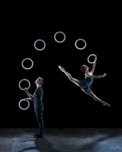 A circus juggler tosses rings in an arch behind him, the first ring is caught by a dancer in a split leap.