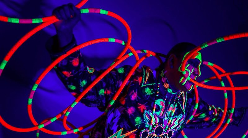 A hula hoop artist spins brightly-colored neon hoops under a black light