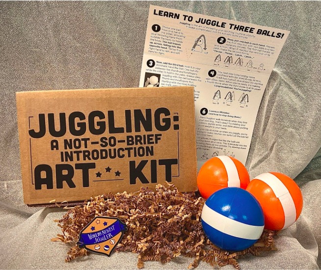 An opened Juggling: A Not-So-Brief Introduction Art Kit, displayed as a box next to blue-and-orange juggling balls and pamphlet