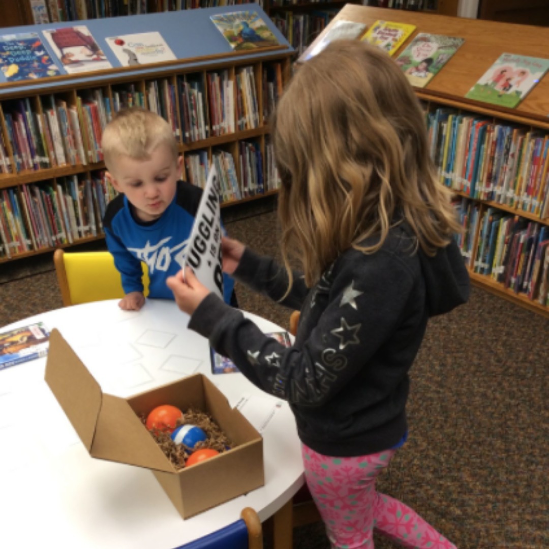 A boy and girl open their juggling kit in front of library shelves