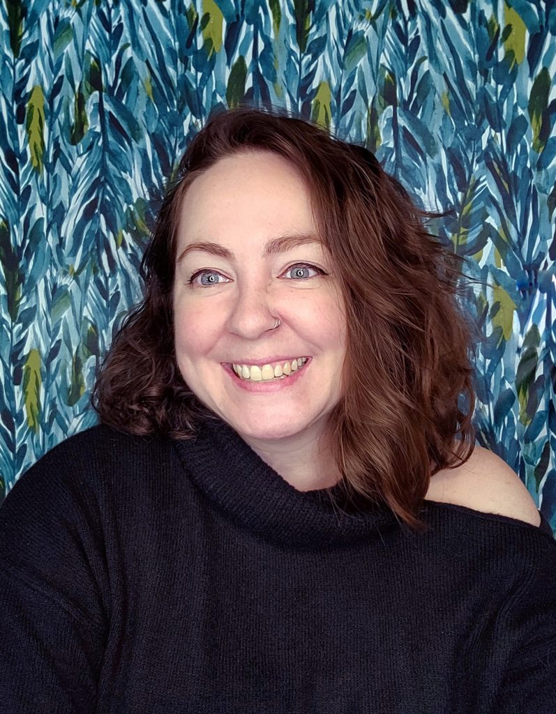 Jenny Leigh Du Puis smiles against a blue green patterned background wearing a black sweater.