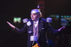 Ukrainian Clown Misha Usov, wearing a tin bowl on his head, juggles pots and pans. He wears a suit with orange pants.