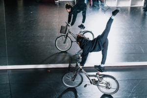 In front of a studio mirror, a Taiwanese female acrobat balances one-legged on her bicycle