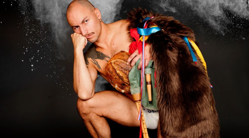 JD Brousse, circus performer, in (le) Pain, his solo show. Acrobat JD kneels on one knee, elbow propped on the other, with colorful props and brown fur pelt draped over his shoulder