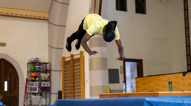 Tumbling practice in Philadelphia's Circus Campus, part of AYCO Festival 2022. A dark-skinned circus student in a yellow shirt practices mini trampoline tumbling over a blue mat