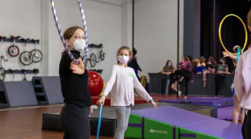 AYCO Festival 2022 hula hoops workshop in the studio of Philadelphia's Circus Campus. Two girls in masks stand catacorner to the blue gymnastics mats. The one with glasses holds a hula hoop that frames her face