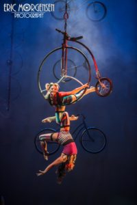 Cirque Mechanics female acrobats hang on a bicycle-like circus apparatus. One acrobat carries the other in the arc of her bent knee