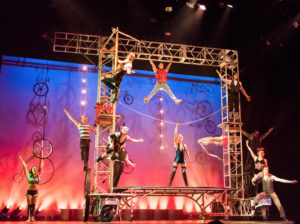 Cirque Mechanics performers hang off, or pose inside, of a metal truss. On the stage wall behind them are some of Chris Lashua-designed circus bikes and unique circus apparatuses.