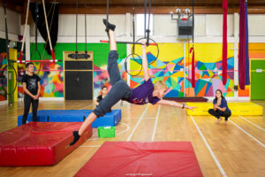 Galway Community Circus training room. A circus student hangs in a back bend from an aerial hoop over a red mat.
