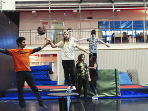 Acrobatics lessons. Circus student teacher, a young man wearing a Zirkus Cirkus t shirt, leads students in an exercise balancing on mats