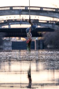 Julia Baccellieri, Black aerialist and circus artist, hangs upside-down from their dance trapeze under Rochester Bridge