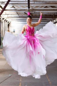 Stephanie Little Thunder Morphet-Tepp, BIPOC American hair hanging artist, suspended from parking garage ceiling beams. She wears a dramatic pink-and-white gown with a long, billowing skirt