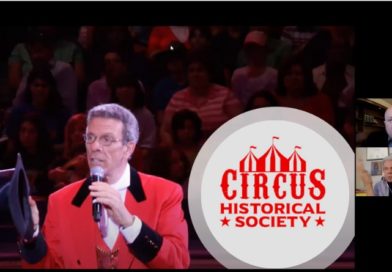 Paul Binder, co-founder of Big Apple Circus, in a ringmaster's suit. Holding a mic, he speaks before an audience. Image contains Circus Historical Society logo and CHS VP Chris Berry