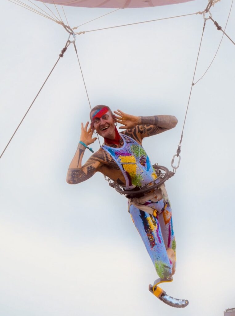 Gerry Tissier, Cirque Bijou Aerialist, hangs from a belted appartus. He puts his hands up by his ears and smiles at the Platinum Jubilee crowd