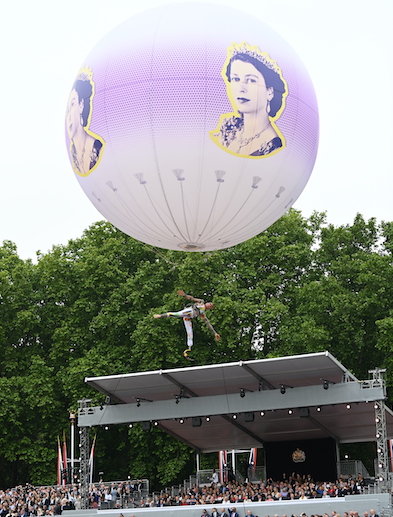 The Queen's Platinum Jubilee balloon, a purple and white balloon with Queen Elizabeth II's face, above an outdoor stage. A Cirque Bijou acrobat does a back kick while standing on the stage roof