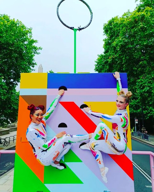 Two Cirque Bijou acrobats, smiling young women in colorful bodysuits, hang from a climbing wall at the Queen's Platinum Jubilee Pageant