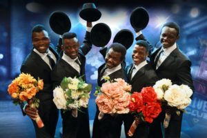 The Black Blues Brothers troupe. Five young Kenyan men in suits hold bouquets of roses and tip their hats to the camera