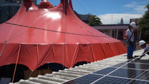 Circo Crescer e Viver tent, a red canvas circus tent, gets rigged with solar panels by two Brazilian men in hard hats