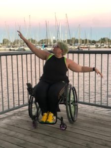 Shay Elrich, a white non-binary person in their mid-30’s with short faded blue hair sits in their manual wheelchair arms outstretched, one above and below their head. The sky behind them is pink, blue and white and the large moon is just above their head through the masts of boats lining the background. They are wearing a black tank top and black track pants with rainbow stripes down the sides. A bright green binder is visible underneath the edges of their tank top.