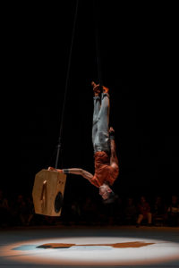 a straps artist in jeans and an orange t-shirt hangs upside down by one foot next to a suspended, large speaker.