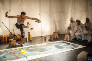 An acrobat balances on one leg with a paintbrush in his mouth. He paints on a canvas in the style of Jackson Pollock