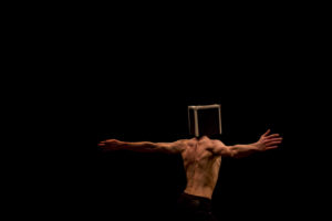 Czech clown Ondřej Holba with a box over his head. He is a young shirtless male performer with his arms outstretched, his bare back to the camera, 