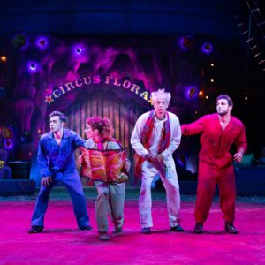 four circus performers look out inquisitively. One is dressed in all blue, another in all red, one in grey, and the fourth in white. The one in grey holds a book or a map.