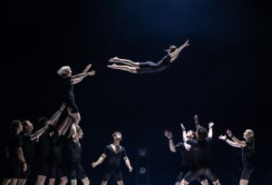 At Edinburgh Fringe Festival 2022, acrobats in black from the Australian company Circa perform in Humans 2.0. Against a black-lit stage backdrop, one acrobat throws another for those in the group beside them to catch