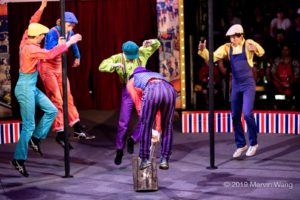 five circus performers in brightly colored hats and shirts and overalls jump in unison. a large hammer is held to the ground.