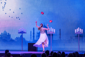 Juggler and meditation teacher Lena Köhn performs diabolo tricks for an audience. Lena is a blonde German woman in a white dress, standing against a stage backdrop with blue castles 