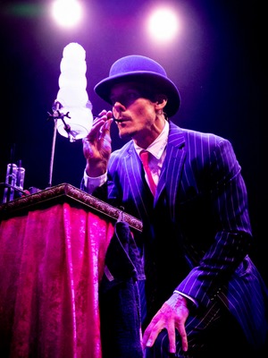 Circus bubble act performer Denis Lock onstage. He wears a pinstriped three-piece suit and bowler hat and blows bubbles out of a pipe or straw